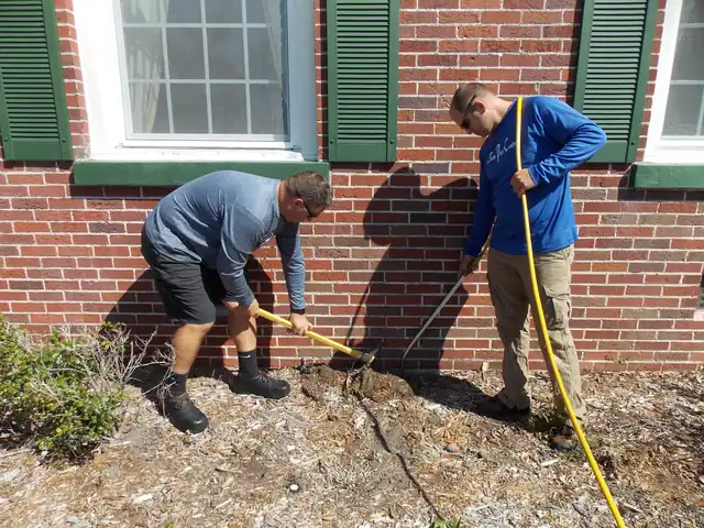 Two technicians trenching along a brick foundation